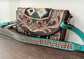 Leather handbag with intricate patterns and a turquoise strap on a wooden surface.